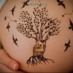 Фото мехенди на животе беременной женщины. Коричневый рисунок с изображением дерева и птиц, выполненный в традиционном стиле.