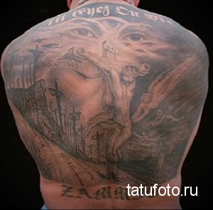 Фото тату на спине с изображением распятия Иисуса Христа. Рисунок выполнен в реалистичном стиле с использованием серых и черных оттенков, с добавлением элементов кладбища и ангельских глаз.