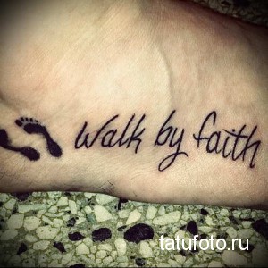 Фото тату на стопе с надписью 'Walk by Faith' и маленькими отпечатками детских ножек, выполненными в черном цвете.