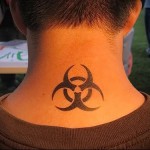 Фото тату: небольшая татуировка с изображением символа биологической опасности на задней части шеи. Рисунок выполнен в черном цвете.