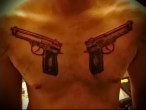 Фото тату двух пистолетов, расположенных симметрично на груди мужчины. Рисунок выполнен в красных и черных тонах.