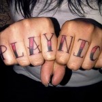 Фото тату: надпись 'PLAY NITCH' на костяшках пальцев рук, выполненная в стиле олдскул с использованием красного и черного цветов.