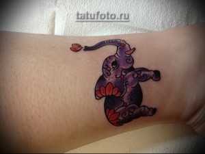 Фото тату слоненка с розовым лотосом на запястье. Нежные пастельные тона, мультяшный стиль, женственная татуировка.