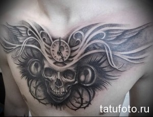 Фото тату на груди мужчины с изображением черепа, на котором расположены часы. Рисунок выполнен в черно-серых тонах с элементами крыльев.
