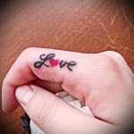 Фото тату: маленькая, черно-красная татуировка надписи 'Love' с изображением сердца на пальце руки. Простой и лаконичный дизайн.