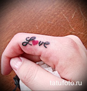 Фото тату: маленькая, черно-красная татуировка надписи 'Love' с изображением сердца на пальце руки. Простой и лаконичный дизайн.