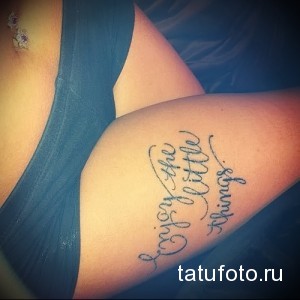 Фото татуировки на бедре с надписью "Enjoy the little things", выполненной каллиграфическим шрифтом. Минимализм, мотивационная татуировка.