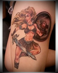 Фото тату на плече с изображением воительницы с мечом и щитом. Рисунок выполнен в ярких цветах с использованием желтого, красного и черного.
