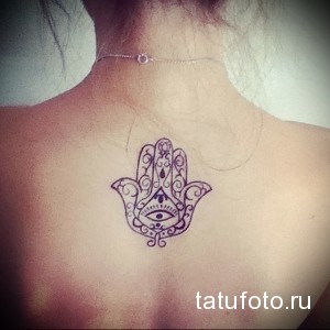 Фото тату Хамсы на спине девушки. Рисунок татуировки в виде руки с открытым глазом, выполненный в фиолетовом цвете, с детальными узорами и орнаментами.