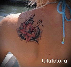 Фото тату красной лилии с черными завитушками на женской лопатке. Детализированный рисунок с мягкими переходами цветов.