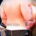 Фото тату надписи 'WICKED' на спине мужчины, выполненной простым шрифтом. Татуировка выглядит немного выцветшей.