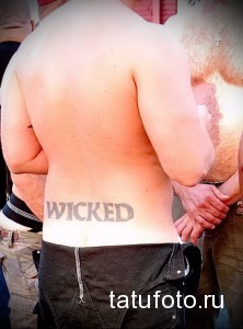 Фото тату надписи 'WICKED' на спине мужчины, выполненной простым шрифтом. Татуировка выглядит немного выцветшей.