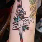 Фото тату на предплечье: кинжал с черепом и надписью 'Owe You Nothing', выполненный в стиле олд скул с использованием ярких цветов и четких линий.
