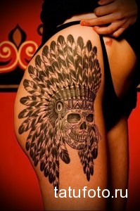 Фото татуировки на бедре в стиле Native American, изображающей череп в головном уборе из перьев, выполненное в черно-белых тонах.
