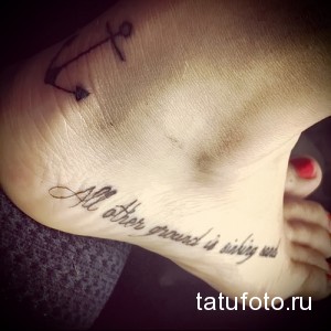 Фото тату на лодыжке с изображением якоря и надписи "All other ground is sinking sand". Минималистичный рисунок татуировки в черном цвете.