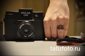 Фотография татуировки в виде маленького фотоаппарата на безымянном пальце руки. Черно-белый рисунок, минималистичный стиль.