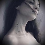 Фото тату на шее и верхней части спины с изображением стаи маленьких птиц, летящих вверх. Черно-белый рисунок татуировки выполнен в графическом стиле.