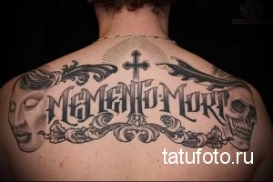 Фото тату готической надписи "Memento Mori" с черепом и масками на спине мужчины. Детализированный рисунок в черно-серых тонах.