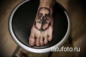 Фото тату черепа с якорем на стопе. Черно-белый рисунок татуировки в стиле олдскул с четкими контурами и традиционными элементами.