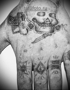 Фото татуировки на груди и руках мужчины: череп с кинжалом, звезды, кресты и другие символы тюремной татуировки. Рисунок выполнен в черном цвете.