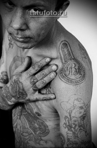 Фото татуировки на плече мужчины: медальон с инициалами, кресты, звезды и другие символы тюремной татуировки. Рисунок выполнен в черном цвете.