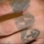 Фотография татуировок на пальцах рук, включая крест в щите, букву 'I' и другие небольшие символы, выполненные в черном цвете.