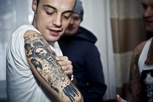 Детализированная черно-серая татуировка на предплечье, изображающая портрет мужчины с крестом и другими религиозными символами.