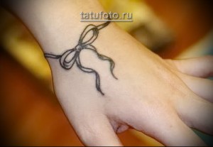 Фото татуировки в виде банта, обернутого вокруг запястья. Рисунок выполнен в тонких черных линиях, создавая изящный и женственный образ.