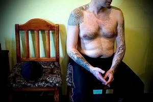 Фотография мужчины с татуировками, покрывающими большую часть его рук и плеч, сидящего на стуле.