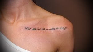 Фото тату: фраза 'What does not kill us makes us stronger' на ключице женщины. Тонкая, изящная надпись, выполненная разными цветами.
