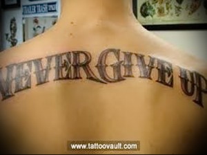 Фото тату надписи 'NEVER GIVE UP' на спине девушки, выполненной черными чернилами в простом, но мотивирующем стиле.