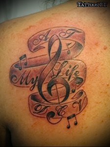 Фото тату на лопатке с изображением скрипичного ключа, нот и надписи 'My Life & My Music'. Черно-серые тона, реалистичный стиль.
