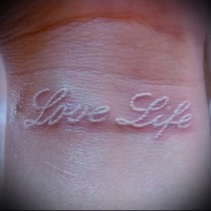Фотография белой татуировки с надписью 'Love Life' на запястье. Простой, но вдохновляющий дизайн, подчеркивающий позитивное отношение к жизни.