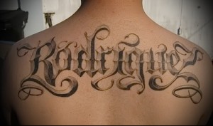 Фотография большой татуировки на спине с именем 'Rodriguez', выполненной в стиле Chicano. Детализированный рисунок с использованием теней и узоров.