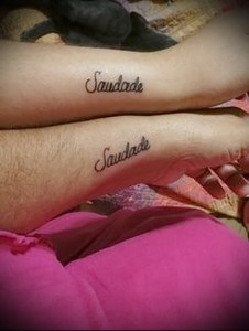 Фото парных тату надписи 'Saudade' на запястьях двух людей, выполненных коричневыми чернилами в изящном шрифте.