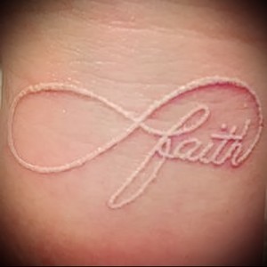 Фотография белой татуировки с надписью 'Faith' в форме бесконечности на запястье. Минималистичный дизайн, нежный и вдохновляющий.