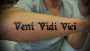 Фото тату латинской фразы 'Veni Vidi Vici' на предплечье мужчины, выполненной черными чернилами в классическом стиле.