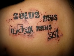 Фото тату надписи «SOUS DEUS IUDEX MENS EST» на плече, выполненной черными чернилами с эффектом потертости. Философская татуировка, выражающая размышления о морали и справедливости.