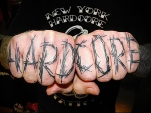 Фото тату на костяшках пальцев с надписью 'HARDCORE', выполненной черными чернилами в стиле графити. Острые углы, брутальный дизайн.