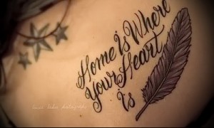 Фото тату надписи 'Home is Where Your Heart Is' и пера на спине, выполненное в черно-серых тонах. Отличный пример для тех, кто ищет вдохновляющие идеи для тату.