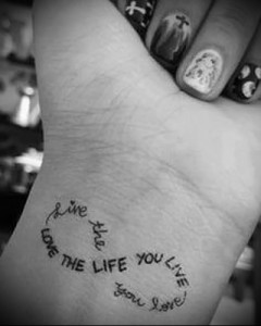 Фото тату надписи 'Live the life you live' на руке, выполненной в минималистичном стиле. Небольшой рисунок татуировки, заключенный в форму сердца.