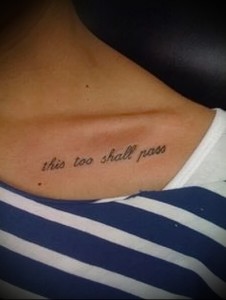 Фото тату с фразой 'this too shall pass', выполненной простым черным шрифтом на ключице девушки. Минималистичный стиль, вдохновляющий смысл.