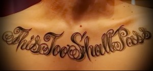 Фото тату с фразой 'This Too Shall Pass', выполненной витиеватым шрифтом с декоративными элементами на груди женщины. Коричневые и черные чернила.