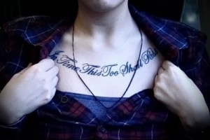 Фото тату с фразой 'In Time This Too Shall Pass', выполненной черным шрифтом на груди девушки. Минималистичный стиль, вдохновляющий смысл.