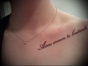 Фото тату с фразой на французском языке 'Aime comme tu l'entends', выполненной изящным шрифтом на ключице девушки. Нежные красные чернила.
