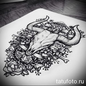 Черно-белое фото тату с изображением черепа быка, украшенного цветами и костями. Детализированный рисунок, готический стиль, символизм.