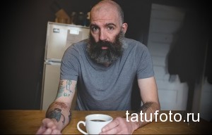 Фото мужчины с длинной бородой и татуировками на руке, держащего чашку кофе. Изображение демонстрирует стиль жизни и любовь к искусству татуировки.