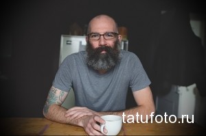 Фото мужчины с бородой и очками, демонстрирующего татуировки на руках и шее, выполненные в различных стилях и цветах.
