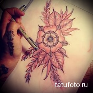 Эскиз татуировки с изображением нежных розовых цветов, выполненный карандашом. Рука мастера с татуировками держит инструмент для рисования.