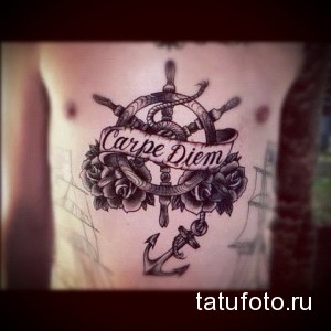Черно-серая татуировка на предплечье с изображением штурвала, обвитого розами, и якоря, с надписью "Carpe Diem".